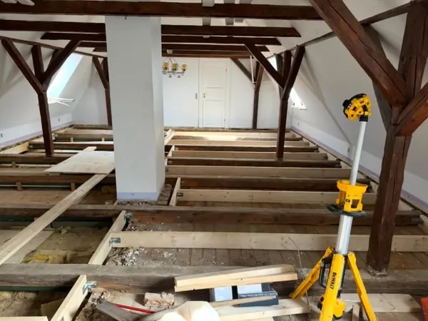 Loft med synlige tagspær og gulvbjælker uden gulvbrædder, med en gul laser på stativ