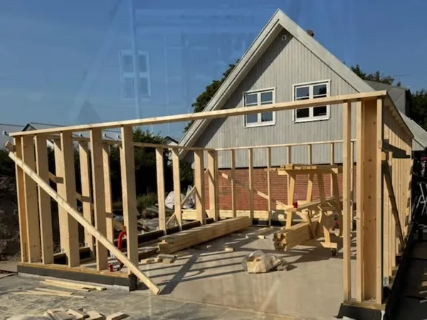 Træskelet af stolper og lægter til en bygning foran et gråt hus med rødt murværk